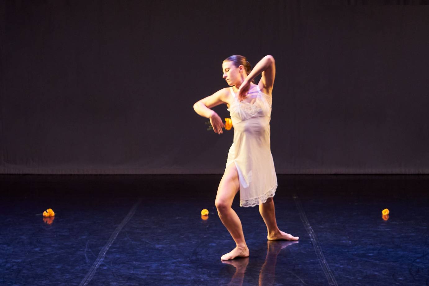 A dancer wearing a white slip lunges forward, head tilted right, elbow lifted.